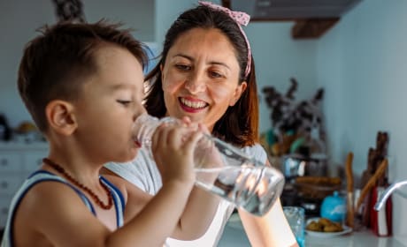 Toddler and mother drink tap water from a glass in a bright, modern kitchen.